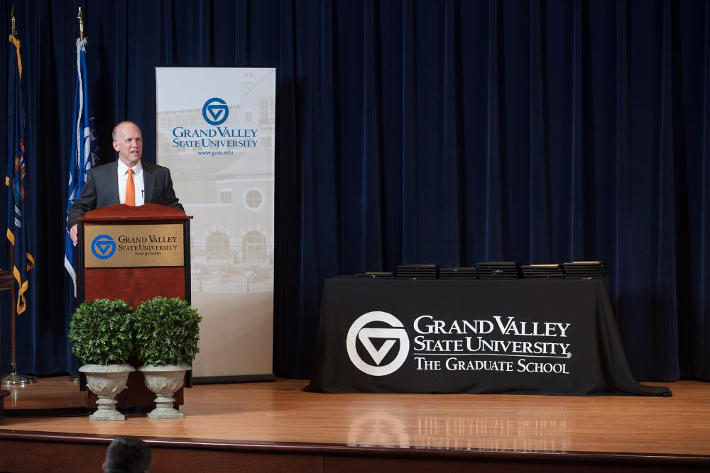 Doctor Potteiger standing on stage at a GVSU podium. There is a table full of awards on stage as well